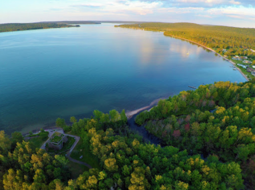 Aerial view of a serene lake surrounded by lush greenery and a sandy shore, with a small structure near the water's edge.