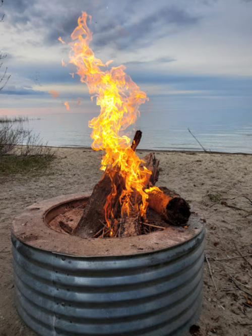 A campfire burns brightly in a metal fire pit on a sandy beach, with calm water and cloudy skies in the background.