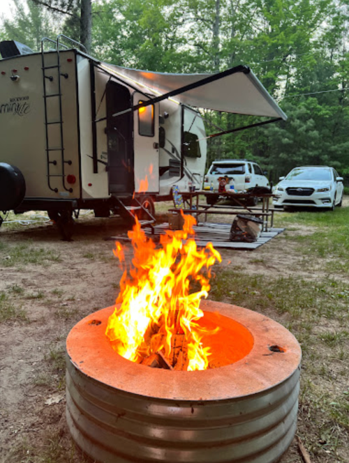 A campfire burns in a metal ring, with a trailer and picnic area in the background surrounded by trees.