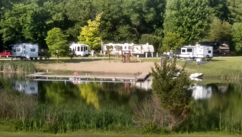 A serene lakeside scene with RVs, a sandy area, playground, and a dock reflecting in the water surrounded by greenery.