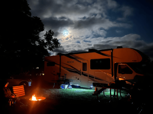 A camper parked at night, illuminated by a campfire and moonlight, with clouds in the sky.