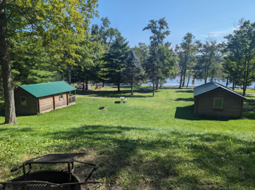 Two wooden cabins on a grassy area by a lake, surrounded by trees under a clear blue sky.