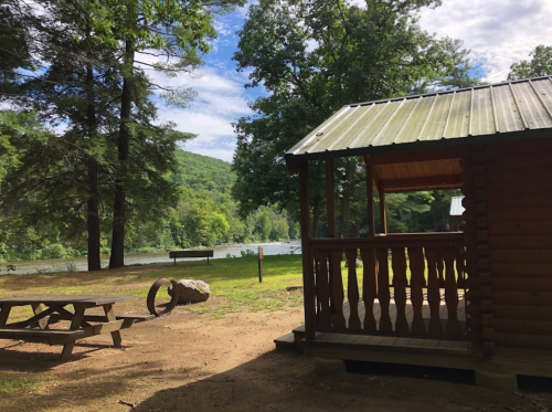 A wooden cabin by a river, surrounded by trees and a picnic table in a serene outdoor setting.