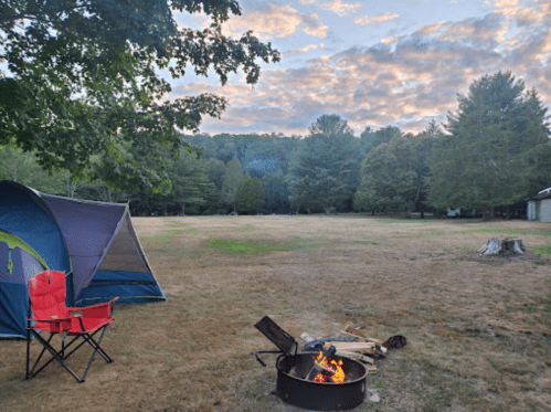 A campsite with a tent, a fire pit with flames, and a red chair, surrounded by trees and a grassy field under a cloudy sky.
