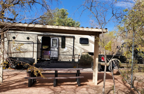 A parked RV under a shelter, surrounded by trees and a gravel area with a picnic table in front.