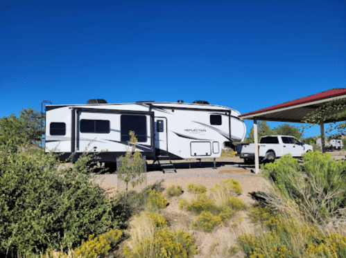A white RV parked under a shelter, surrounded by desert vegetation and a clear blue sky.