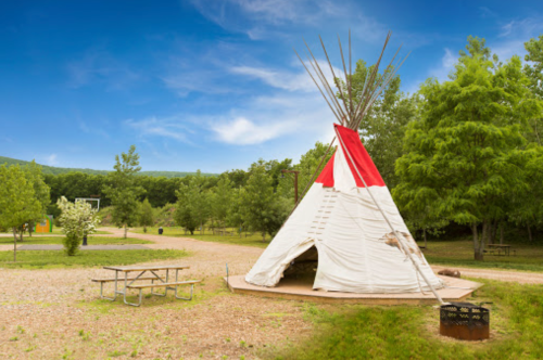 A teepee stands in a grassy area surrounded by trees, with a clear blue sky above and a picnic table nearby.
