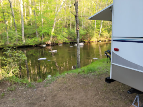 A serene campsite by a calm stream, surrounded by lush green trees and a parked RV.
