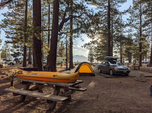 A campsite with a yellow kayak, picnic table, tent, and a car surrounded by tall trees and mountains in the background.