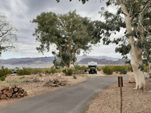 A scenic view of a road leading to a campsite by a lake, surrounded by trees and mountains in the background.