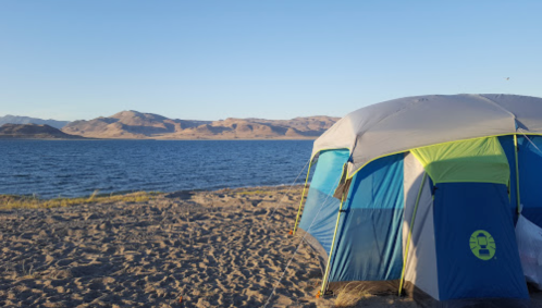 A colorful tent set up on a sandy beach by a lake, with mountains in the background under a clear blue sky.