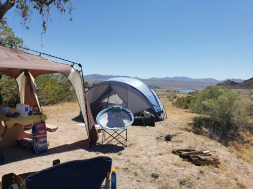 A campsite with two tents, a folding chair, and a scenic view of mountains and a river under a clear blue sky.