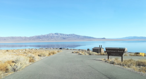 A road leads to Sportsman's Beach campground, with mountains and a calm lake in the background under a clear blue sky.