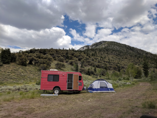 A red camper parked beside a blue tent in a scenic outdoor setting with mountains and trees in the background.