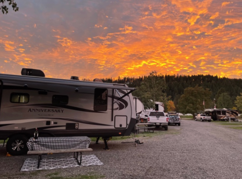 A scenic campsite at sunset, featuring an RV, picnic table, and vibrant orange and pink clouds in the sky.