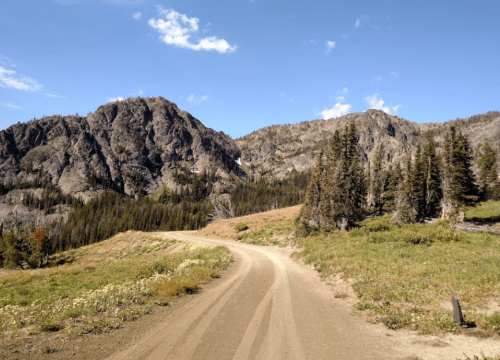 A winding dirt road leads through a mountainous landscape with trees and blue skies.
