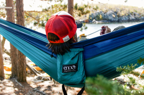 A person relaxes in a blue hammock, wearing a red cap, surrounded by trees and a scenic view of water.