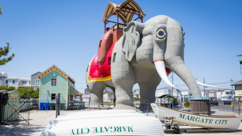 A large, colorful elephant statue with a wooden structure on its back, surrounded by boats and houses in Margate City.