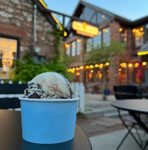 A cup of cookies and cream ice cream sits on a table, with a charming building and string lights in the background.