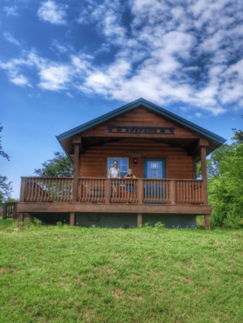 A wooden cabin with a porch, surrounded by greenery and a blue sky, featuring a person standing at the entrance.