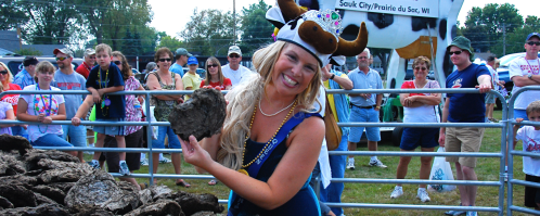A smiling woman in a cow-themed hat holds a large object, surrounded by a crowd at an outdoor event.