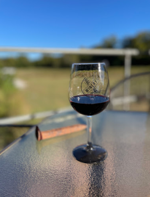 A glass of red wine sits on a table with a winery logo, overlooking a sunny outdoor landscape.