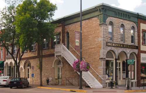 Historic Scriver Building with a stone facade, white staircase, and hanging flower baskets in a charming downtown setting.