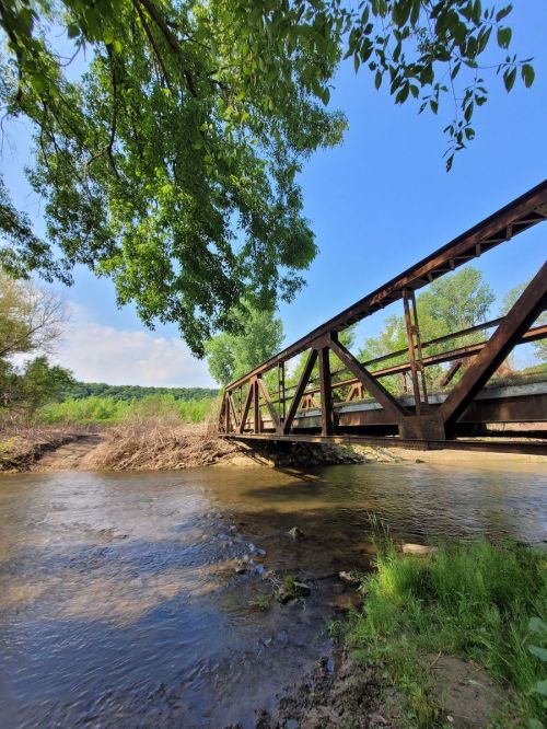 A metal bridge spans a calm stream, surrounded by lush greenery and a clear blue sky.