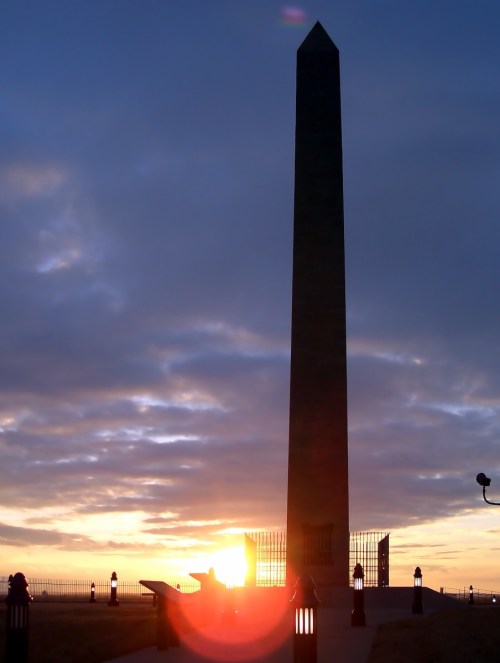 Silhouette of an obelisk at sunset, with clouds and a glowing horizon in the background.