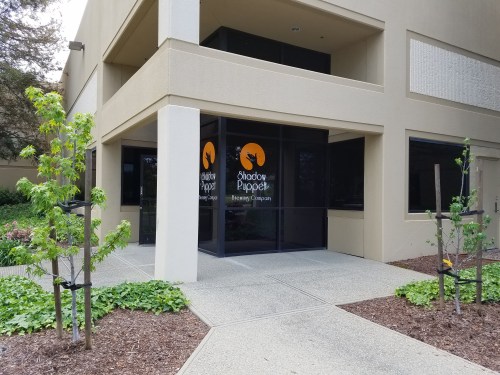 Entrance of Shadow Puppet Brewing Company, featuring large glass doors and greenery around the pathway.