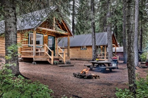 A cozy log cabin surrounded by trees, with a picnic table and a fire pit in a serene forest setting.