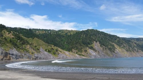A serene beach scene with gentle waves, surrounded by lush green hills under a blue sky with scattered clouds.