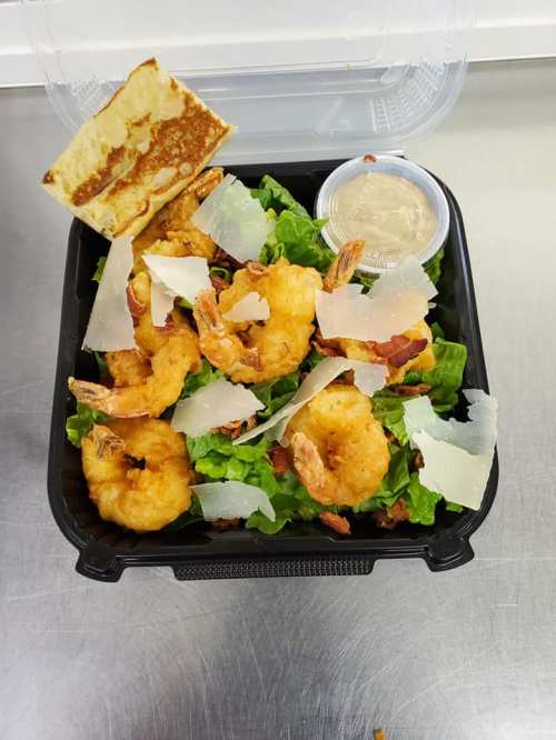 A black takeout container with a salad topped with fried shrimp, lettuce, Parmesan cheese, and a side of dipping sauce.