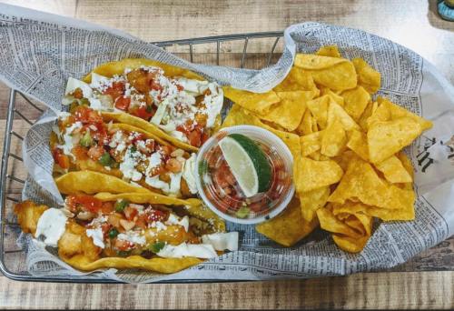 A basket of tacos topped with salsa and cheese, served with tortilla chips and a side of lime wedges.