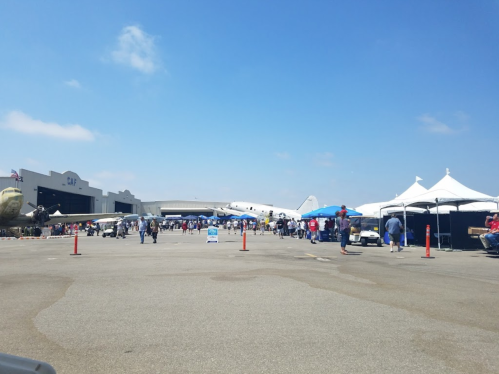 A busy airshow scene with people, tents, and vintage aircraft in the background under a clear blue sky.