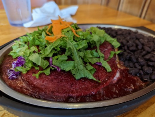 A sizzling plate of red enchiladas topped with fresh greens and carrots, served with black beans on the side.