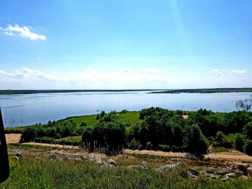 A serene view of a lake surrounded by greenery under a clear blue sky with a few clouds.