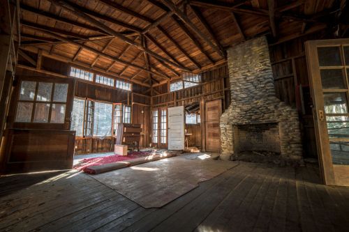Interior of a rustic cabin with wooden beams, a stone fireplace, and large windows letting in natural light.
