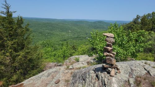 A stone cairn stands on a rocky outcrop, overlooking a lush green valley and distant blue hills under a clear sky.