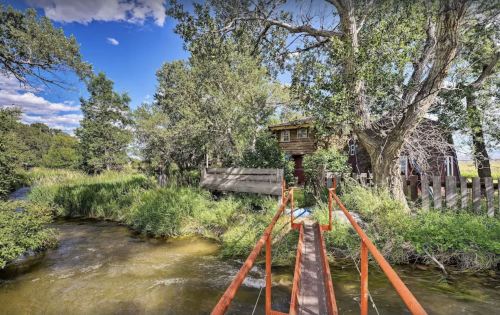 A rustic wooden house by a stream, surrounded by lush greenery and trees, with a small bridge leading to it.