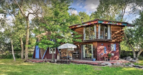 A unique, colorful two-story house surrounded by trees and greenery, featuring a deck and outdoor seating area.