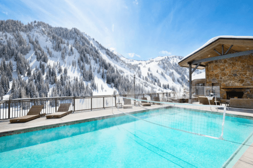 A scenic outdoor pool with a mountain backdrop, surrounded by lounge chairs under a clear blue sky.