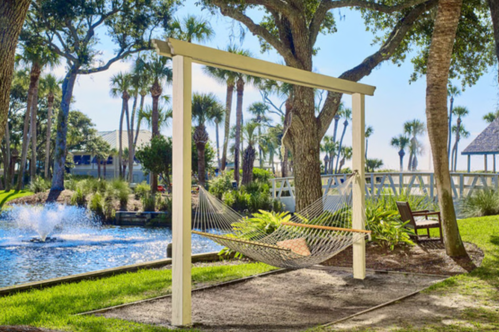 A hammock hangs between two trees by a pond, surrounded by lush greenery and palm trees under a clear blue sky.