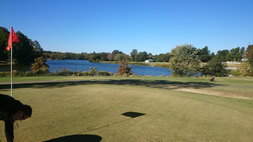 A golf course with a red flag on the green, overlooking a serene lake and trees under a clear blue sky.