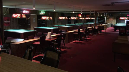 A dimly lit pool hall with several green tables, bar seating, and illuminated signs along the walls.