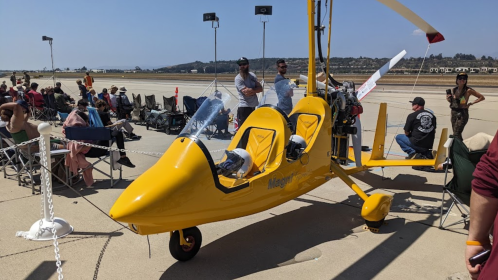 A bright yellow ultralight aircraft on a runway, with spectators seated nearby at an airshow.