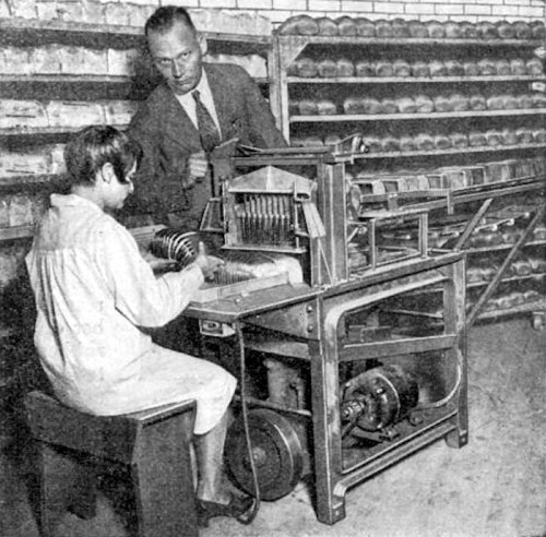A man supervises a child operating an early mechanical device in a workshop filled with shelves of products.
