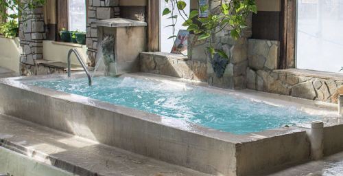A serene indoor hot tub with bubbling water, surrounded by stone walls and greenery. Natural light filters in through windows.
