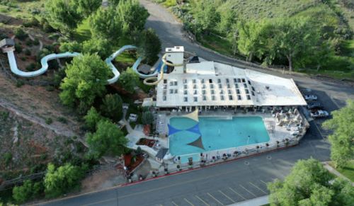Aerial view of a pool area with a water slide, surrounded by greenery and a parking lot nearby.