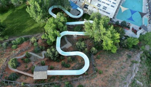 Aerial view of a winding water slide surrounded by trees, with a pool and buildings in the background.